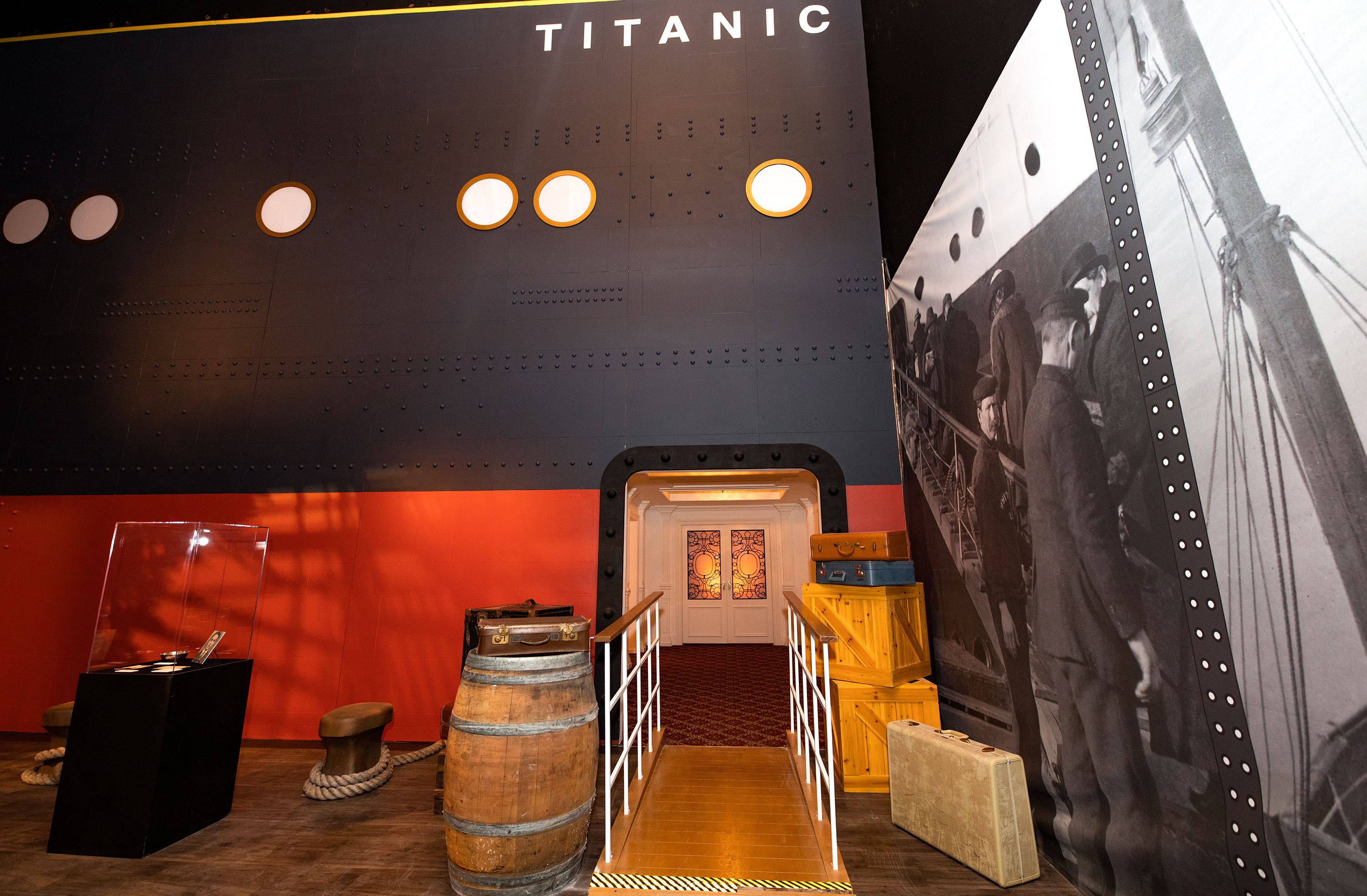 Dark riveted wall with orange-ring portholes and the title "TITANIC." Red lower wall, barrel with rope, crates, and railing to a doorway; right side shows a grayscale mural of sailors.