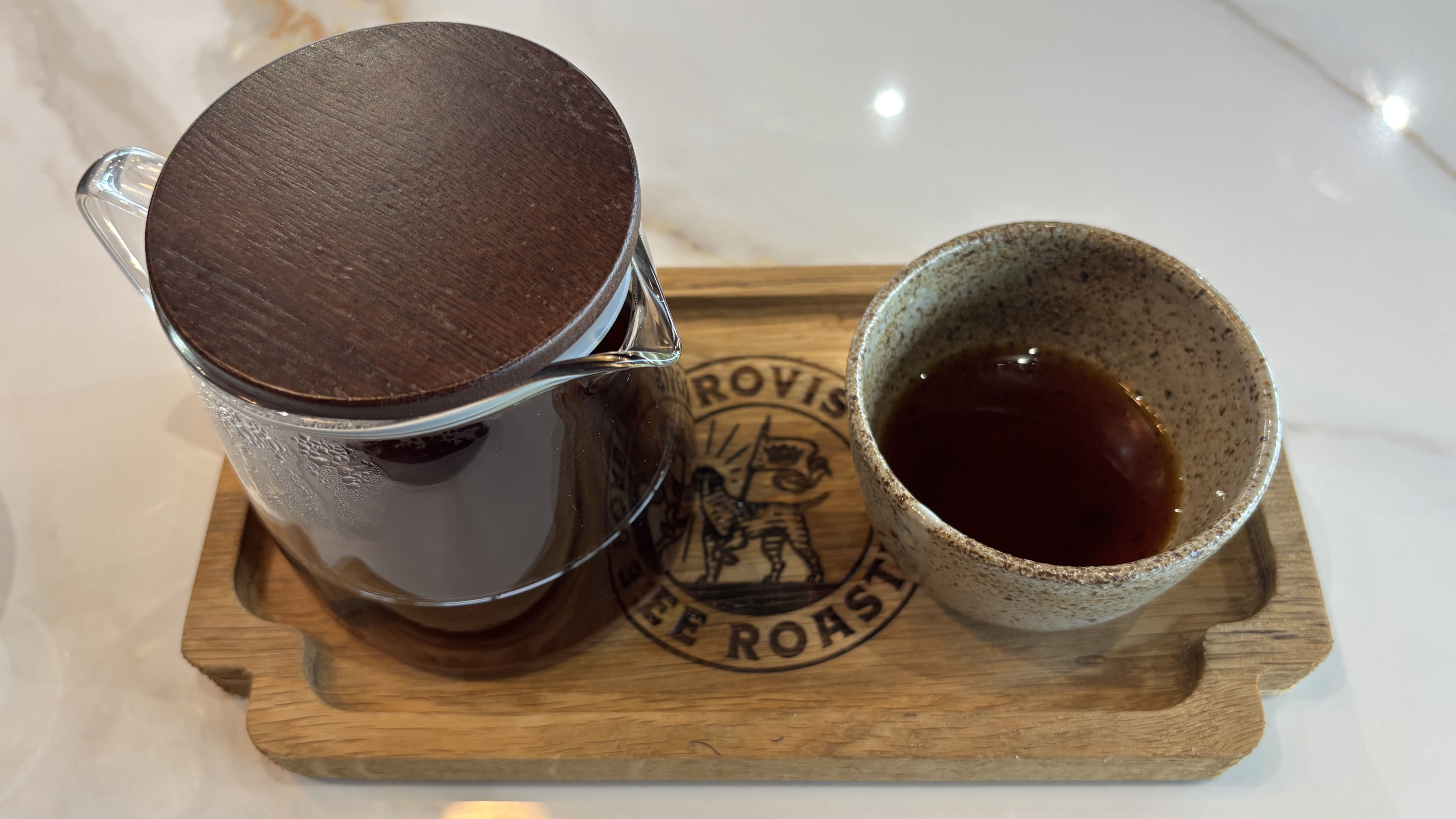 A glass coffee carafe with a round dark wooden lid sits on a wooden tray beside a speckled ceramic mug filled with dark coffee on a light marble counter.