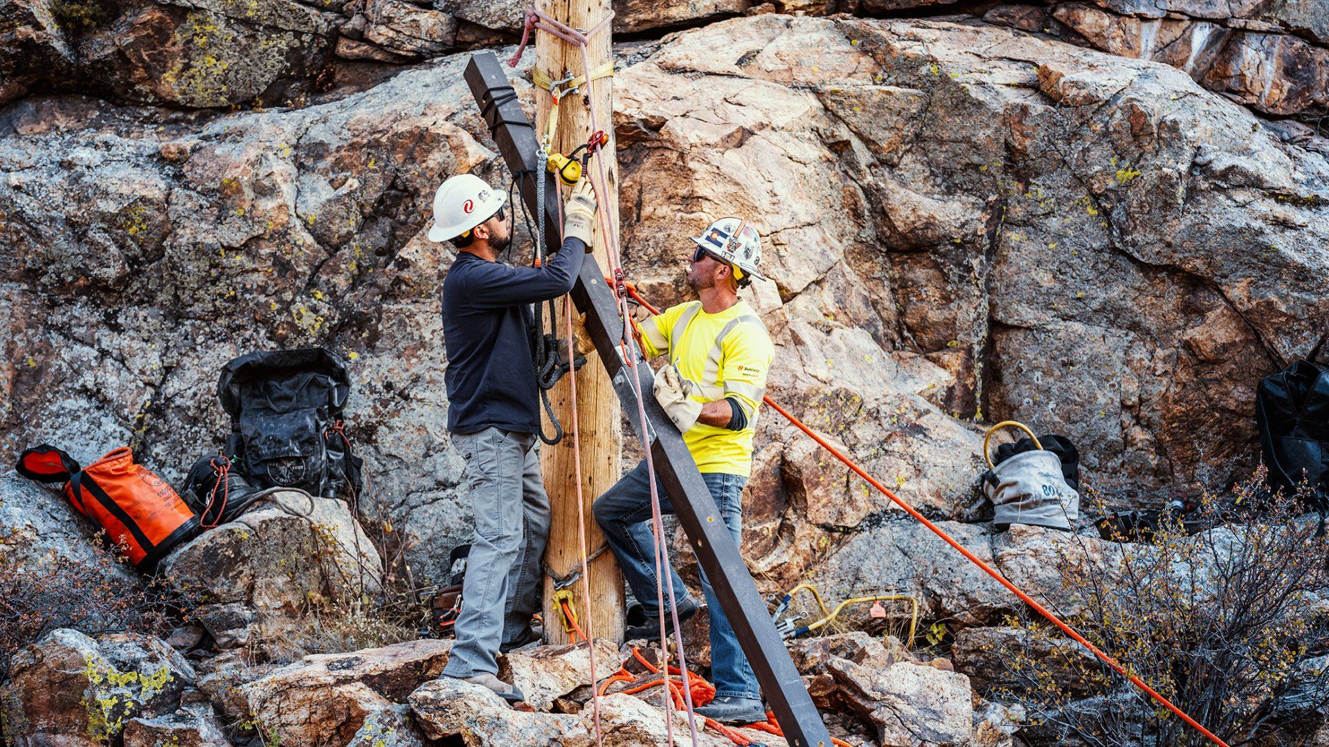 Two construction workers wearing helmets and gloves work on securing equipment to a wooden pole amid rocky terrain with tools and bags scattered around.