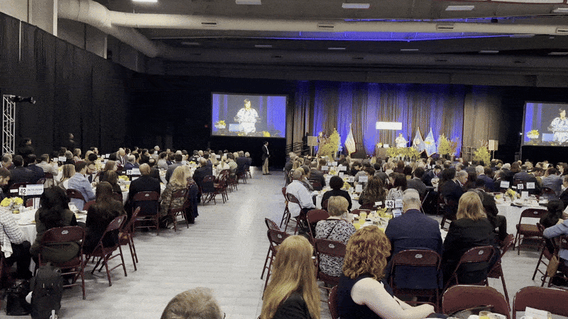 Large formal event in a dark hall with rows of round tables and people seated facing a stage with a speaker displayed on two large screens against blue curtains.