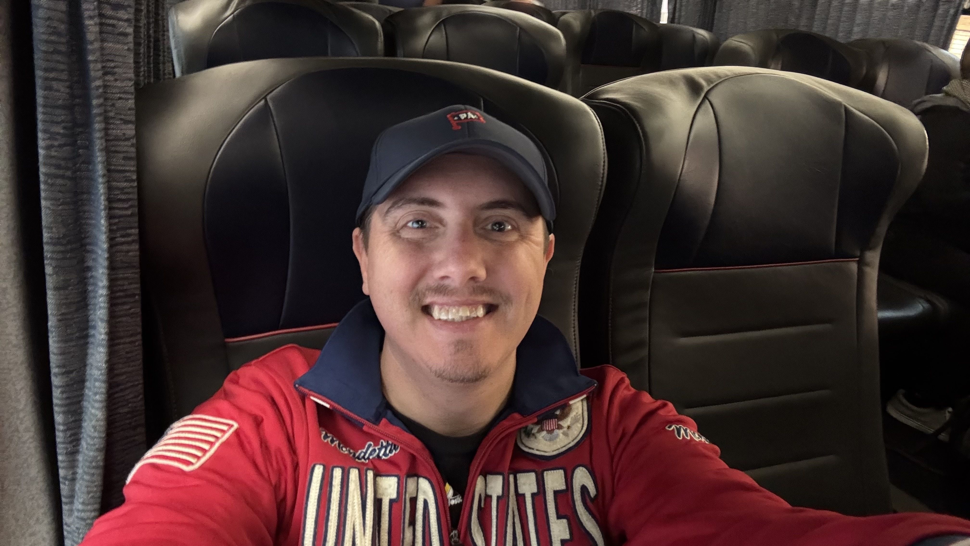 Smiling man wearing a navy cap and red jacket with "United States" text, sitting on a black leather bus seat with more similar seats in background.