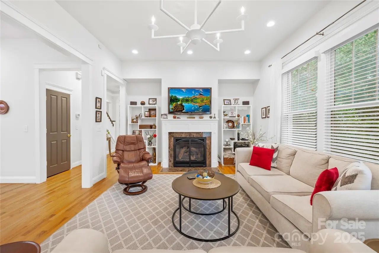 Bright living room with white walls, wood floors, beige sofa with red pillows, brown leather chair, round coffee table, fireplace, TV above it, and large windows with white blinds.