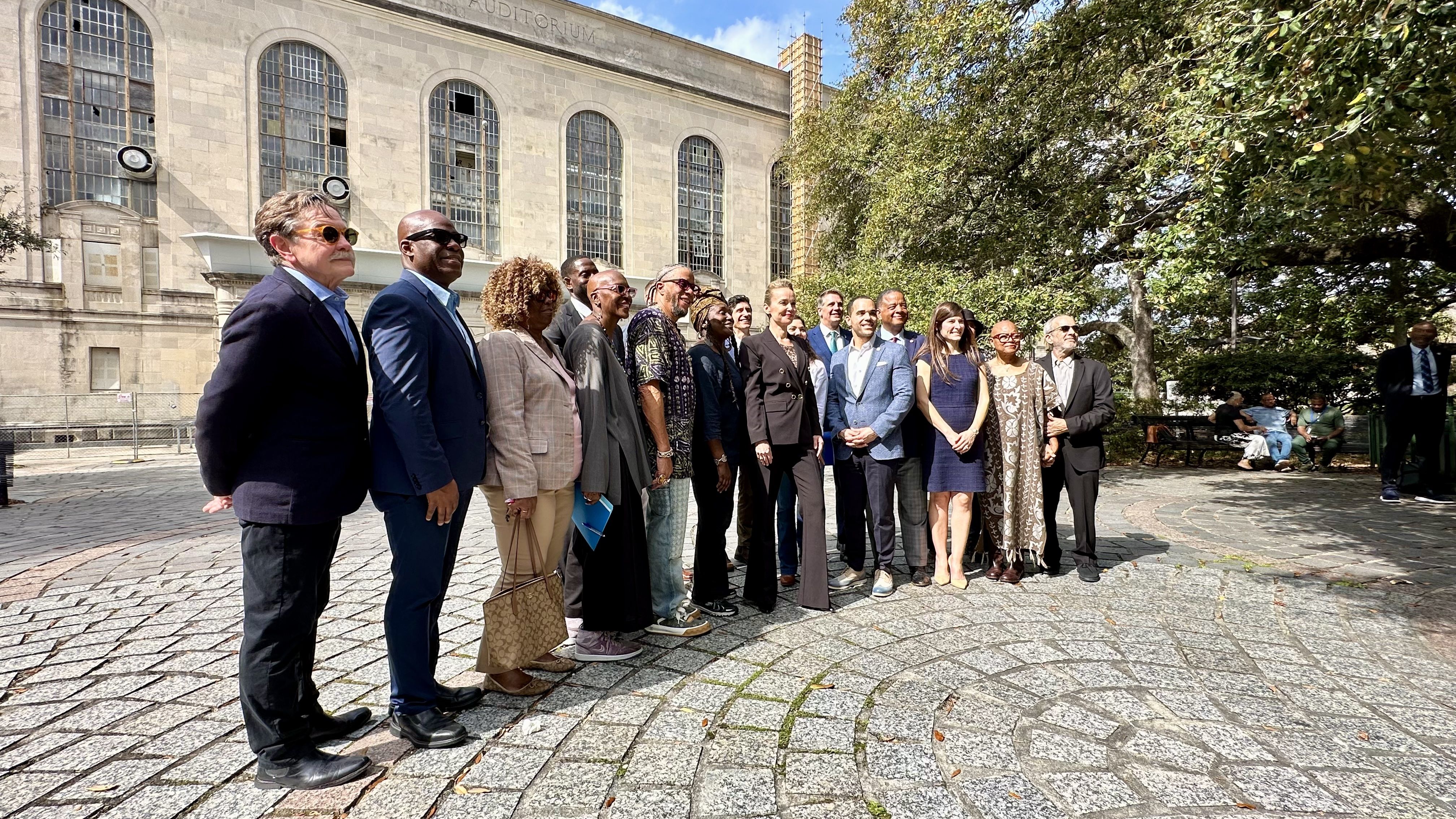 Group of diverse people dressed in business and casual attire standing on a patterned stone plaza with a large building and trees in the background on a sunny day.