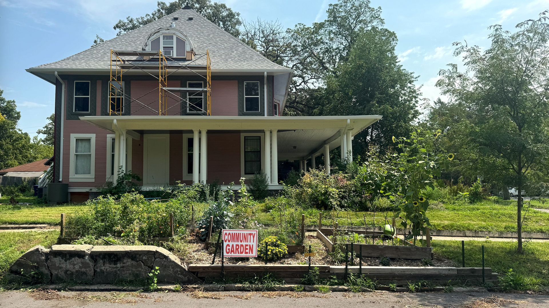 Two-story pink house with a gray roof and a rounded attic window, yellow scaffolding on the front, white porch columns, and a community garden in the front yard under a blue sky.