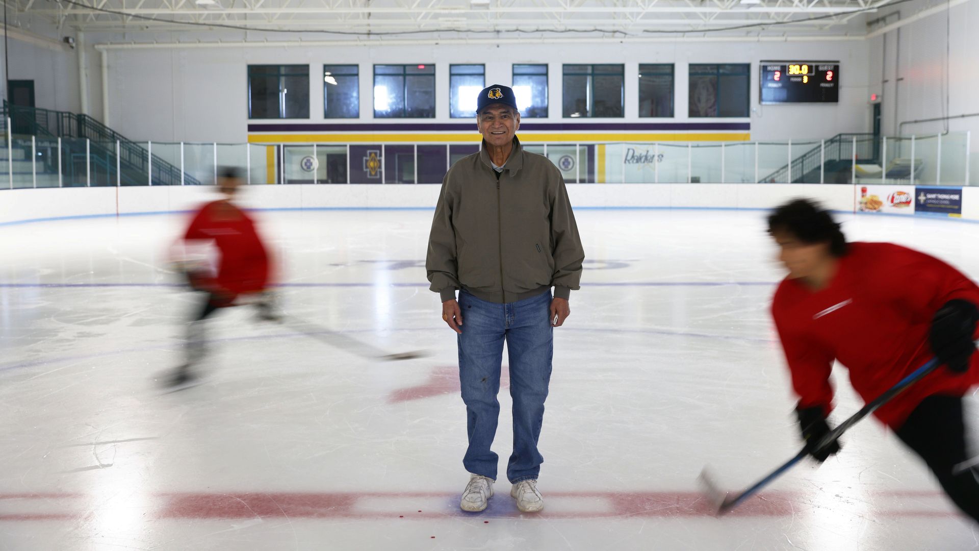 An older man with brown skin in a khaki jacket and a hat with an Indian head logo on it stands and smiles for a photograph in the center of an ice rink as the blurred figures of two younger hockey players skate circles around him.