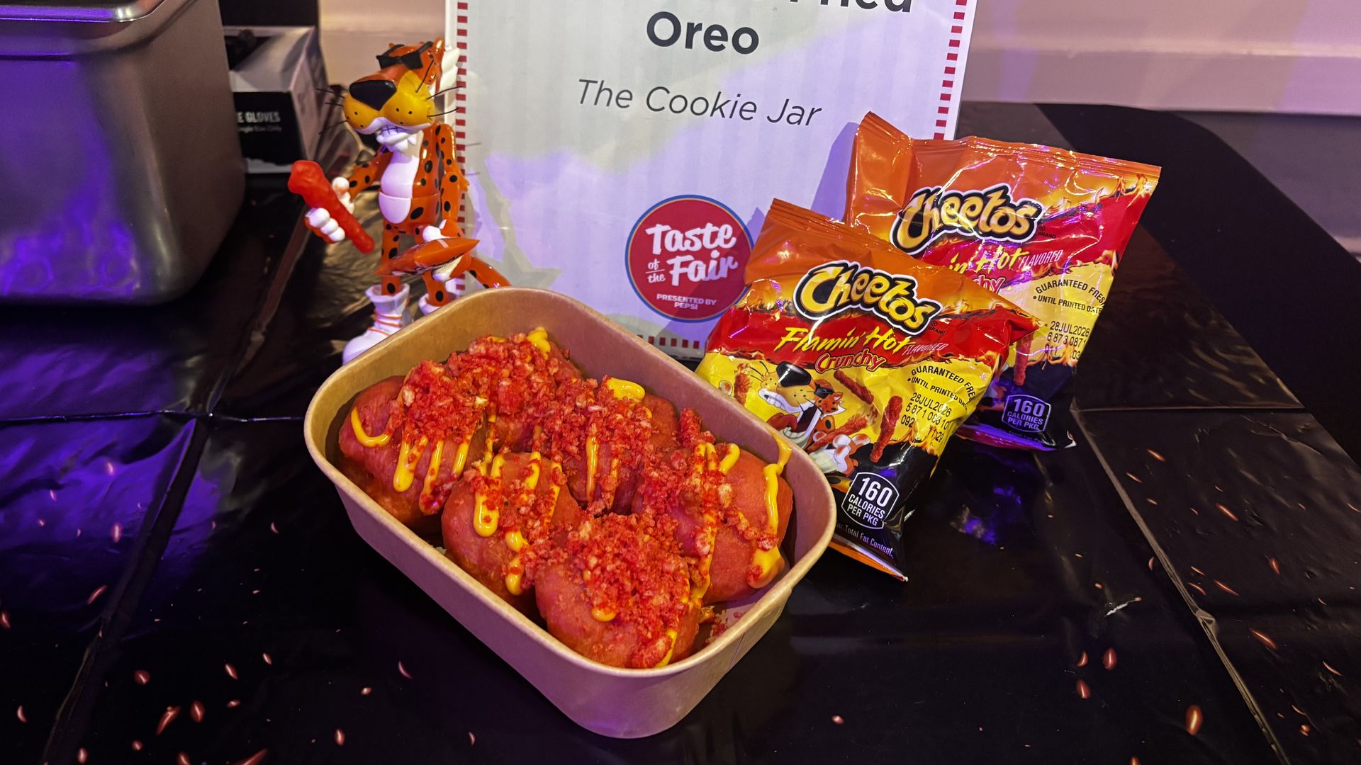 Tray of hot dogs topped with mustard and red crumb on a black counter, beside orange Cheetos bags. A leopard plush stands near a sign that says \"The Cookie Jar\" and \"Taste of the Fair.\"