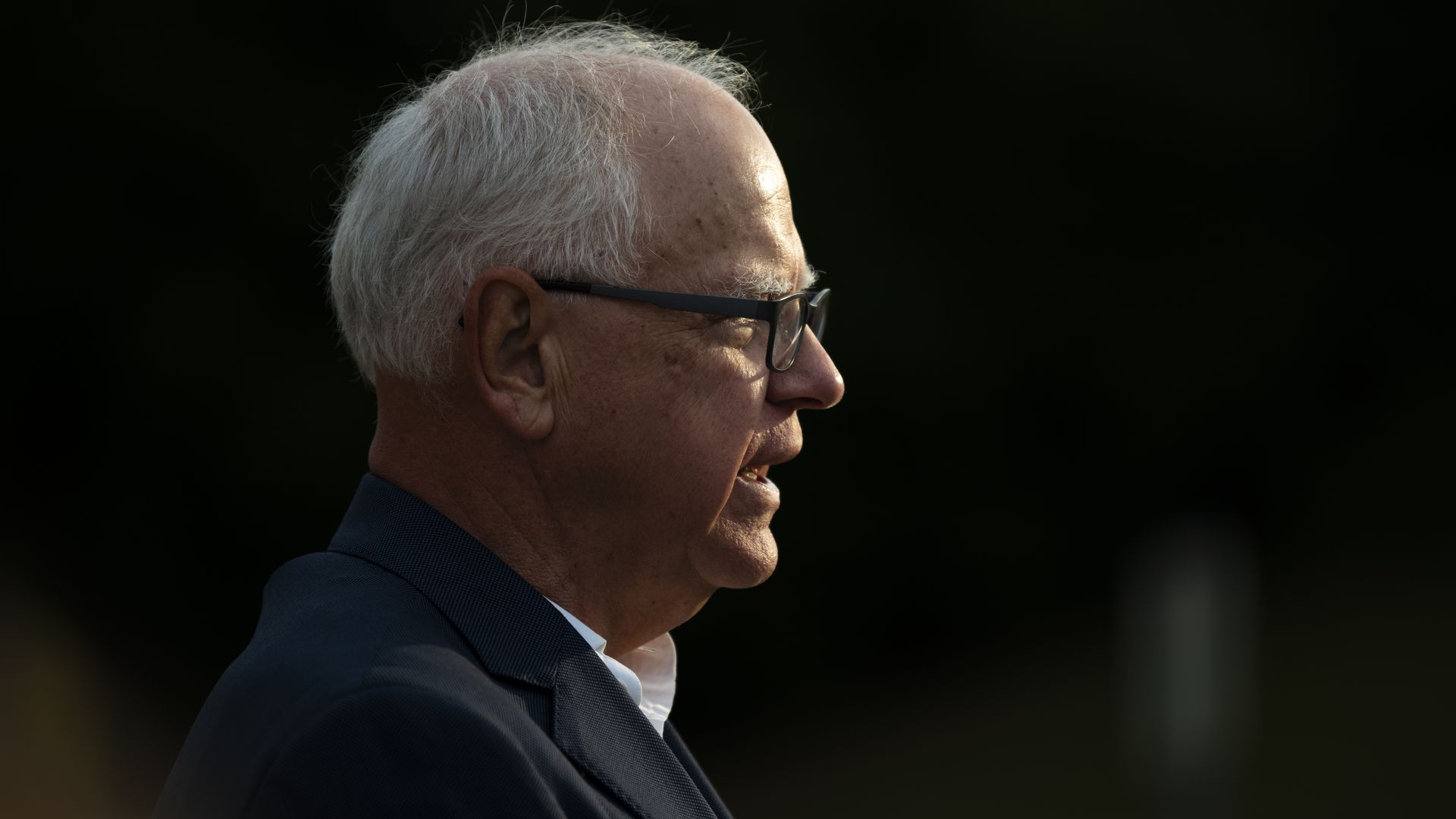 Profile of a man with white hair and glasses, wearing a dark jacket and white shirt, against a dark background with soft lighting on his face.
