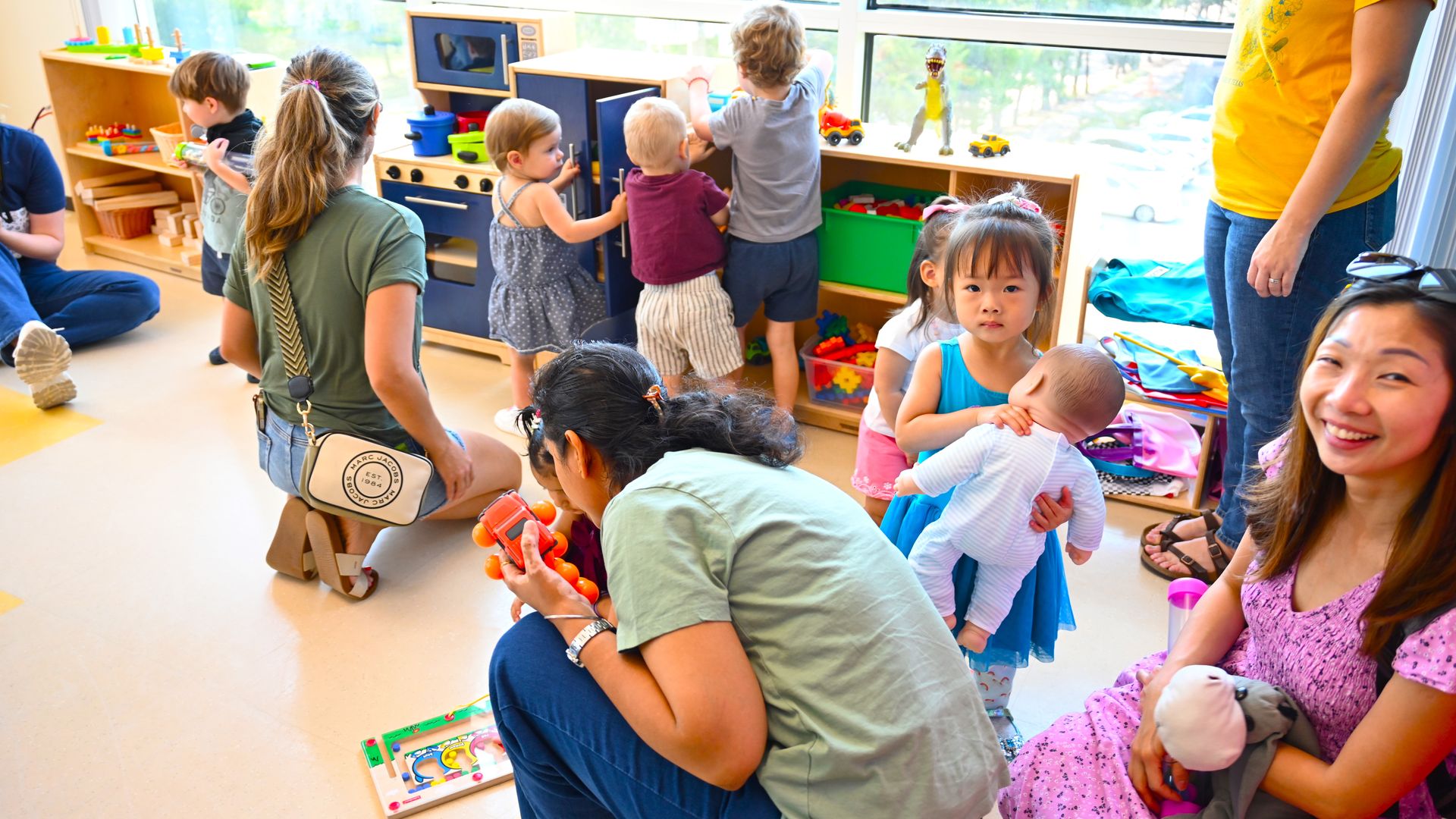 Children and adults playing in a bright room with toys and wooden shelves. A girl in a blue dress holds a baby doll, and a smiling woman in a purple dress holds stuffed toys.