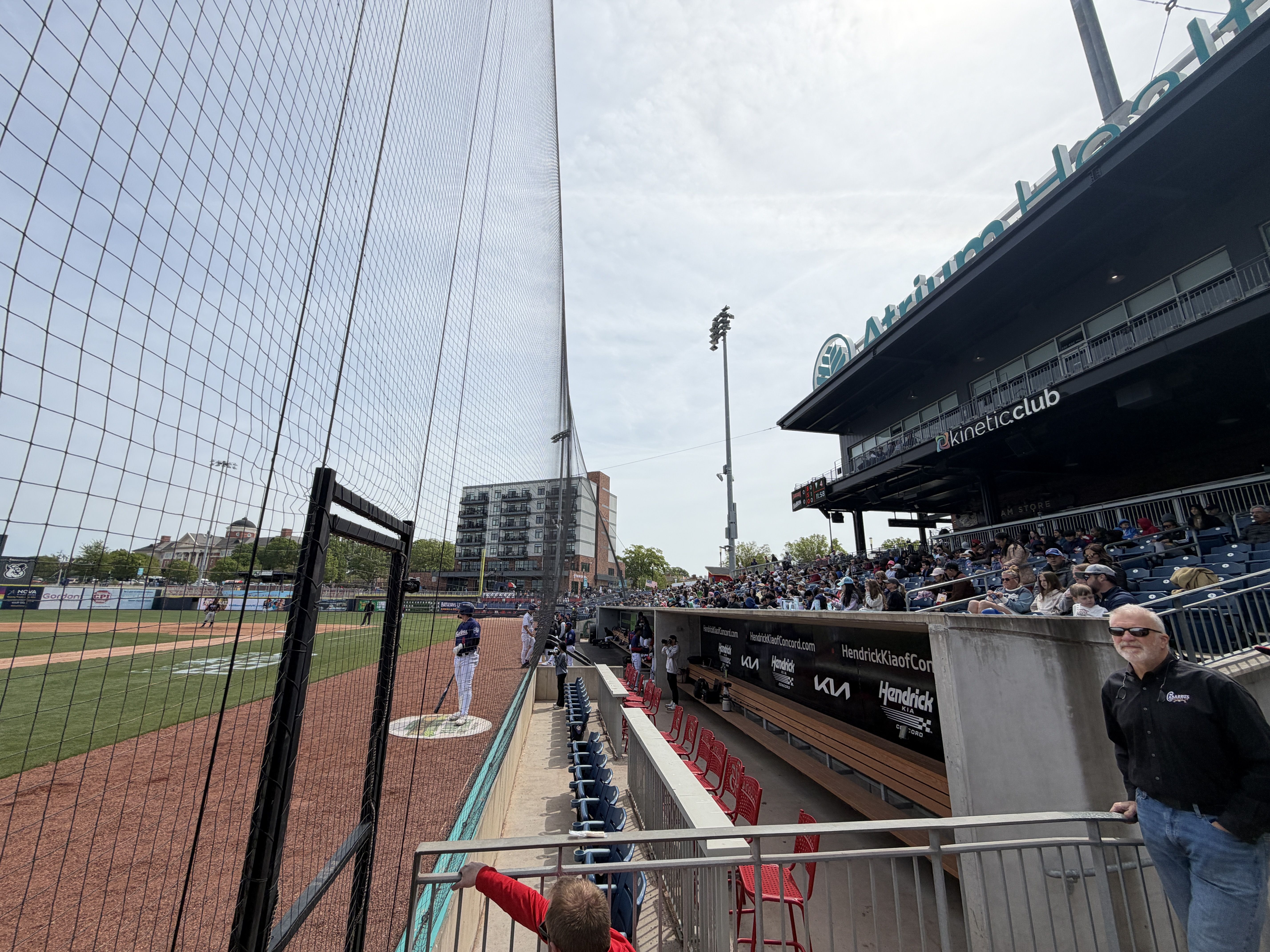 View from behind the net at a sunny baseball game. A pitcher on the mound faces home plate as a batter stands nearby. Fans fill the right-field stands; a man in a black shirt watches from the railing.