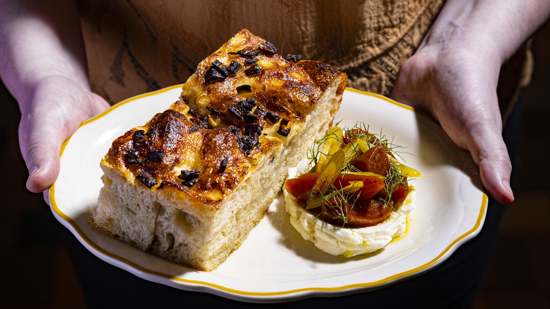 A person holds a white plate with a yellow rim, presenting a thick, golden-brown loaf topped with dark crispy bits. Beside it, a dollop of whipped cheese is topped with tomato slices and dill.