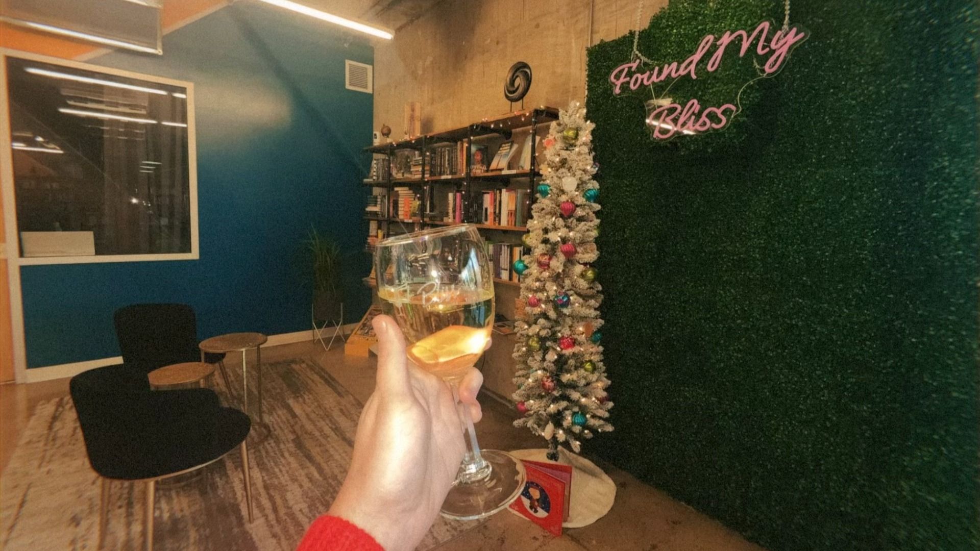 Hand holding a glass of white wine indoors near a decorated white Christmas tree with colorful ornaments and a green wall displaying pink neon sign "Found My Bliss."