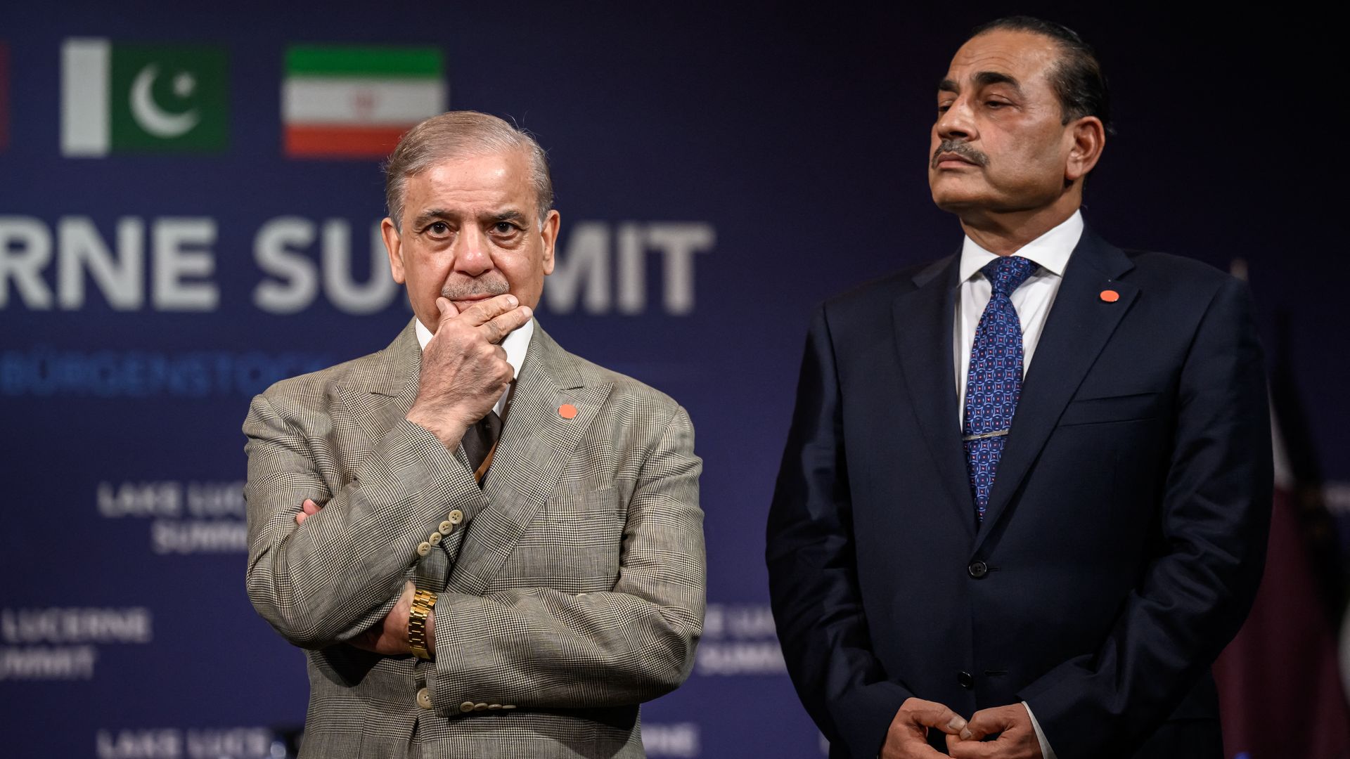 Two men in suits on a stage with a dark blue backdrop and flags; left man in a tan checkered suit with a hand near his mouth, right man in a dark suit with a blue patterned tie.