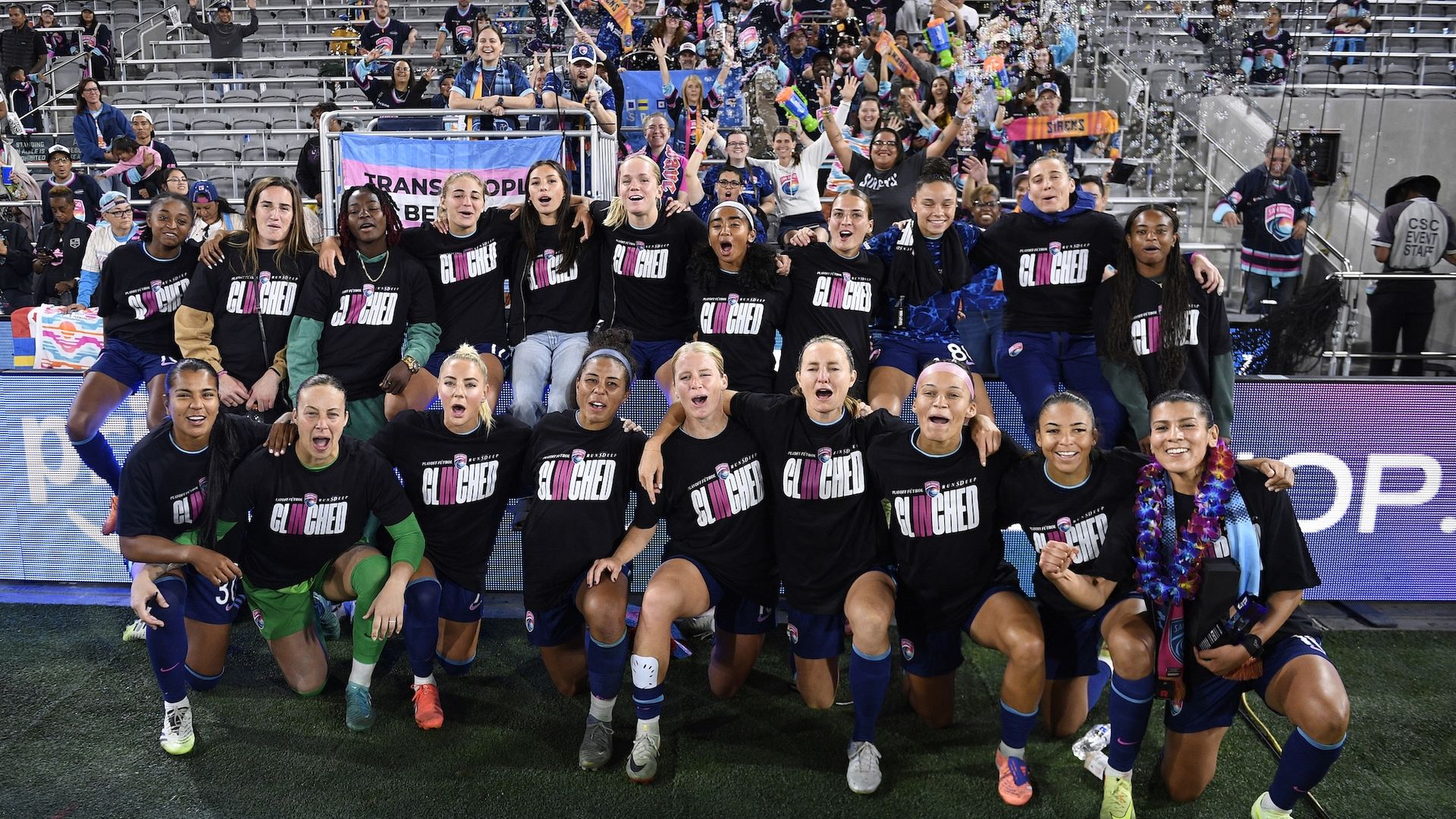 San Diego Wave players, wearing black tee shirts with white and pink writing that says "CLINCHED,"cheer while posing for a photo with fans in the stands after a win.