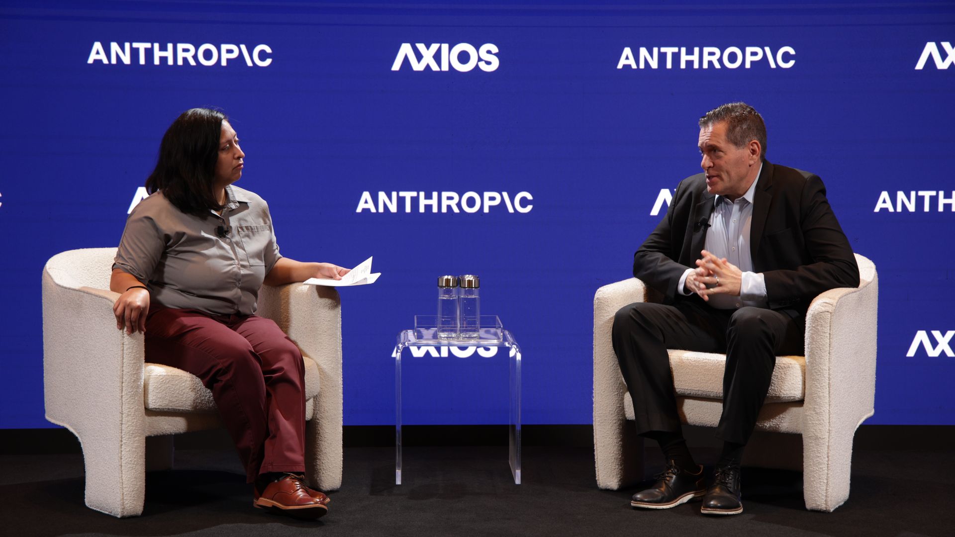 Panel discussion with a woman on the left and a man on the right, seated in cream chairs on a blue backdrop with "ANTHROPIC" and "AXIOS" logos; a glass table with water bottles sits between them.