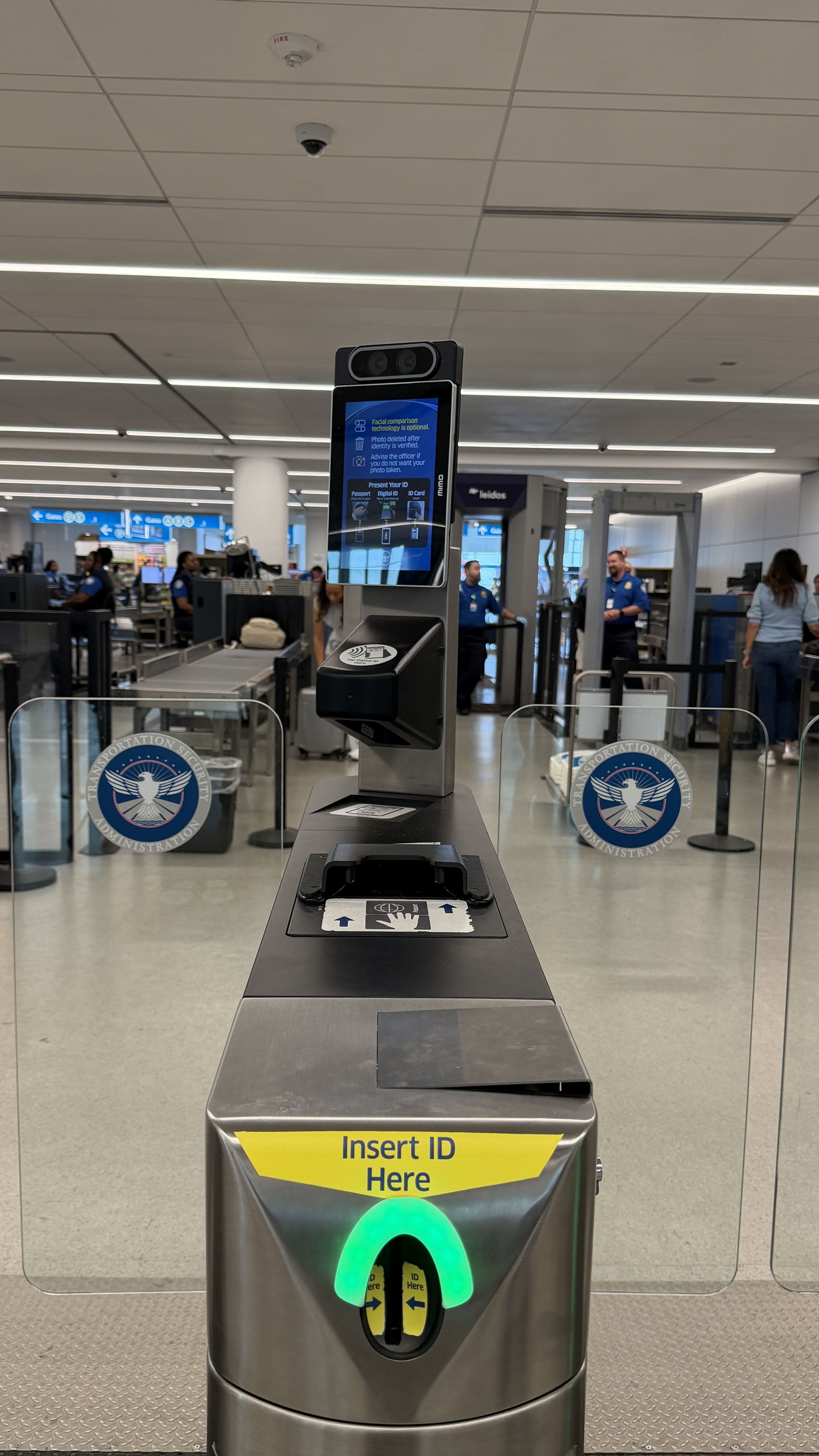 Airport security checkpoint with a tall metal ID scanner, blue display screen and top camera, glass barriers with TSA logos, and a yellow Insert ID Here sign; a green indicator glows.