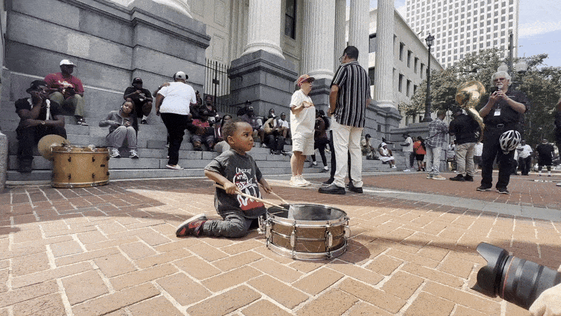 Young boy kneeling on brick ground playing a snare drum with drumsticks in front of a crowd gathered near a stone building with columns during daytime.