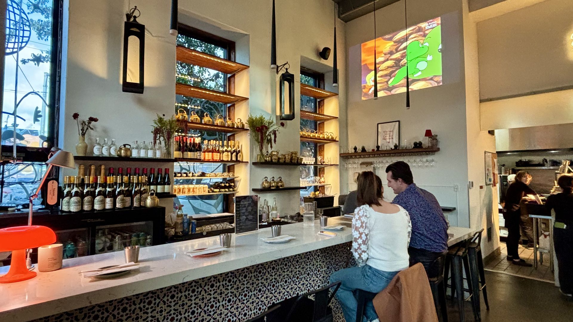 Cozy bar with patterned counter, bottles on shelves, two people sitting at the bar, and a colorful cartoon projected on the white wall above, warm lighting enhancing the space.