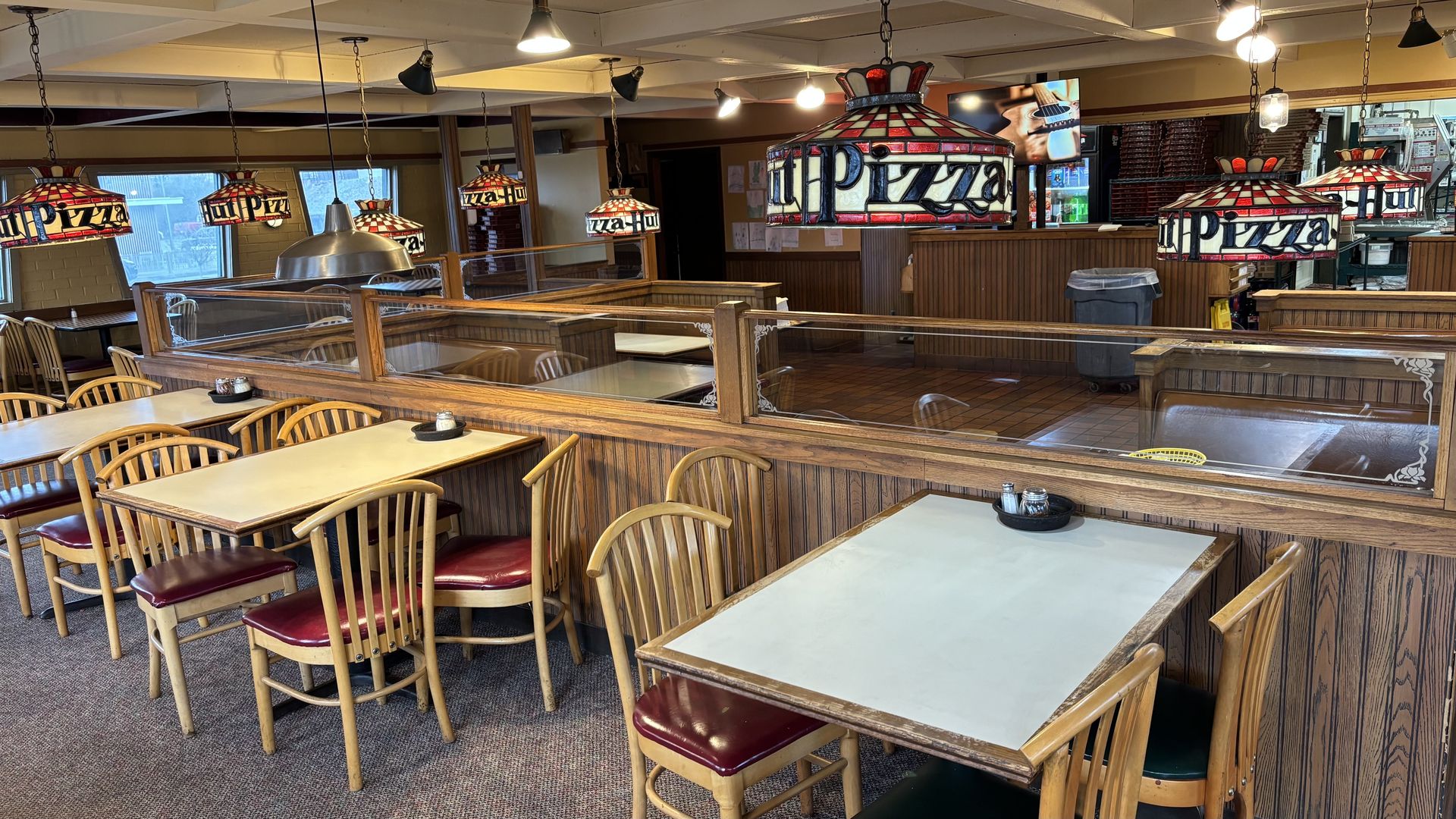 Interior of a casual pizza restaurant: wooden booths and tables, red-cushioned chairs, glass dividers, and stained-glass pizza lamps hanging from the ceiling; kitchen and trash can visible in back.