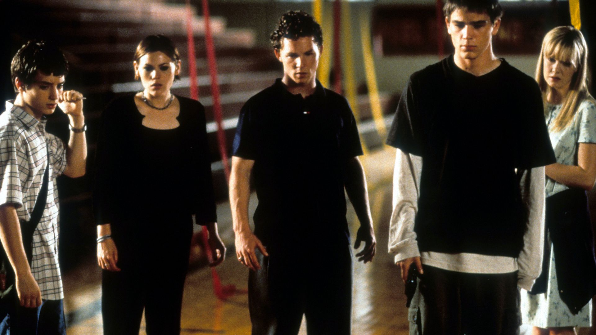 A group of teenagers stand together in a gymnasium looking at something ominously. 