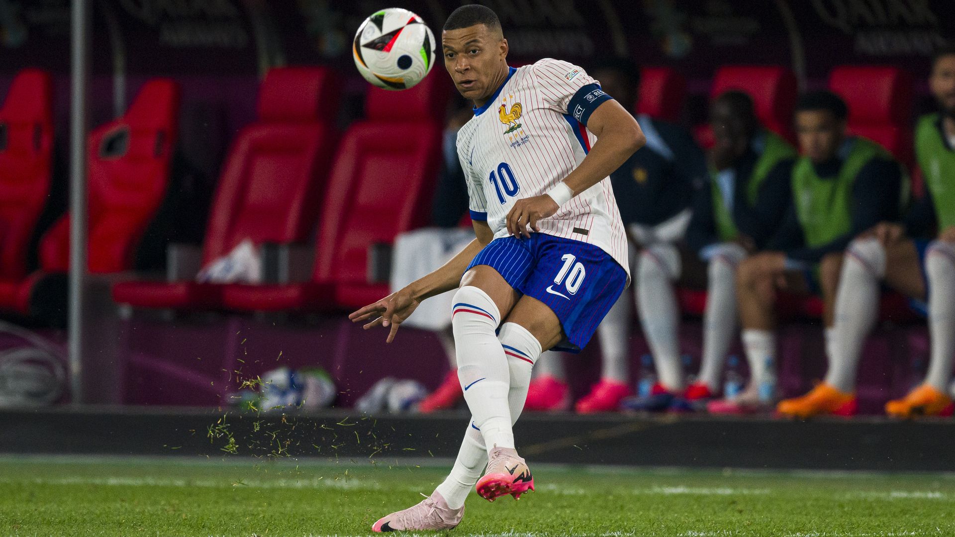 A man with short-cut hair wearing short sleeves, shorts and high socks kicks a ball in the air while on grass in an arena. 