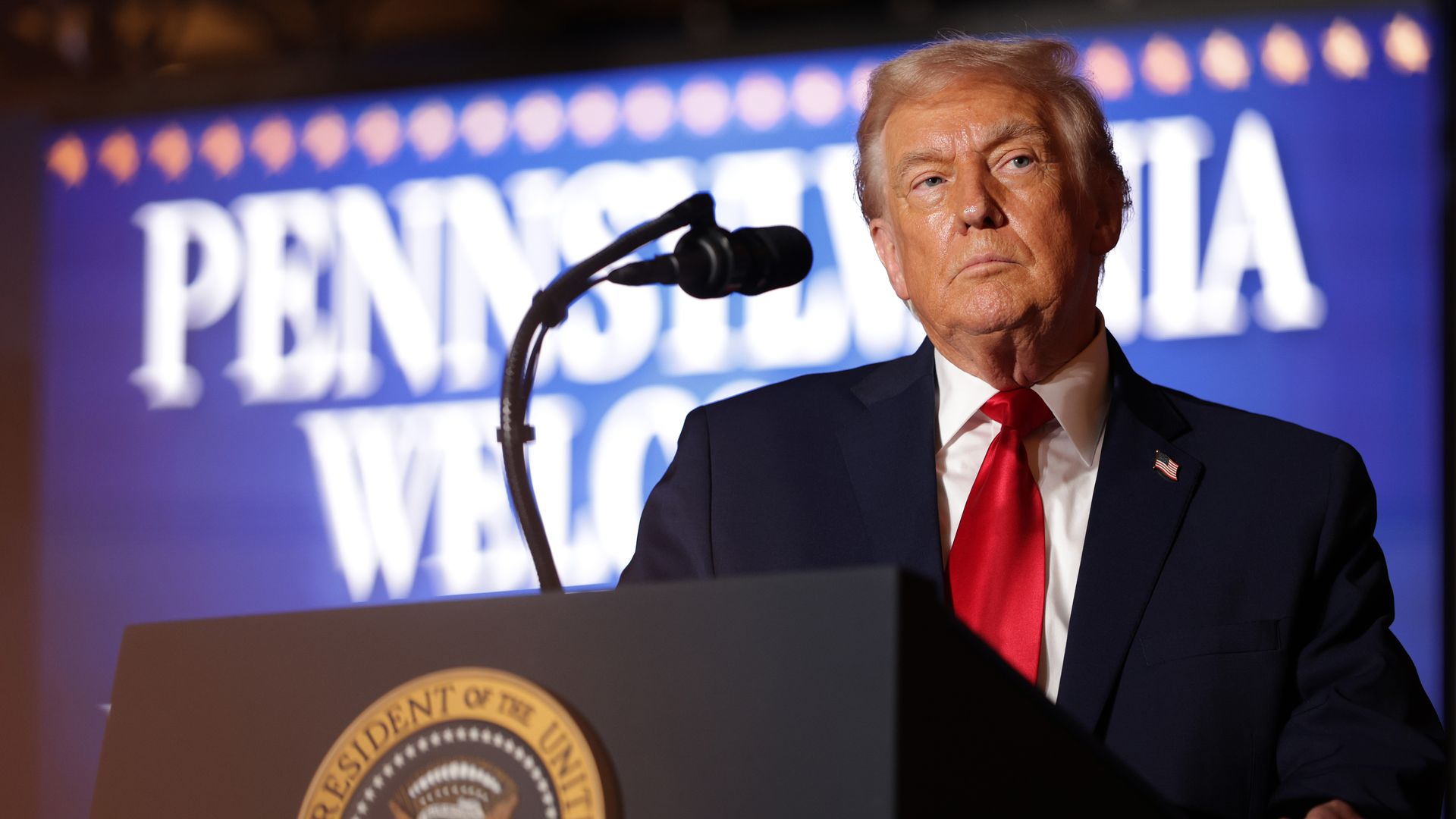 Trump wearing a blue suit jacket, white shirt and red tie behind a podium 