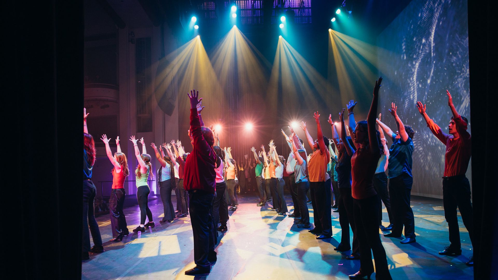 Wide stage scene with many performers raising arms in colorful shirts beneath vivid blue, yellow and teal spotlights; dark curtains frame the sides as bright lights cast long shadows on the floor.