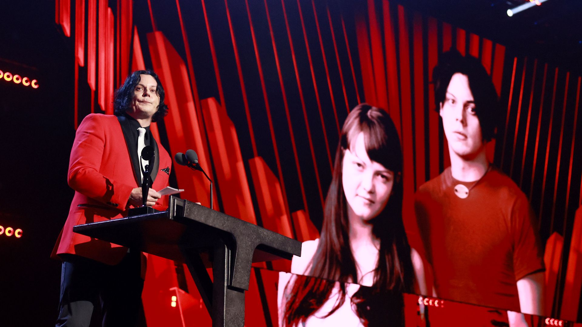 Person in red jacket and black tie speaking at podium with two microphones, large screen behind showing black-haired man and woman with long hair in red and white tops against red and black background.