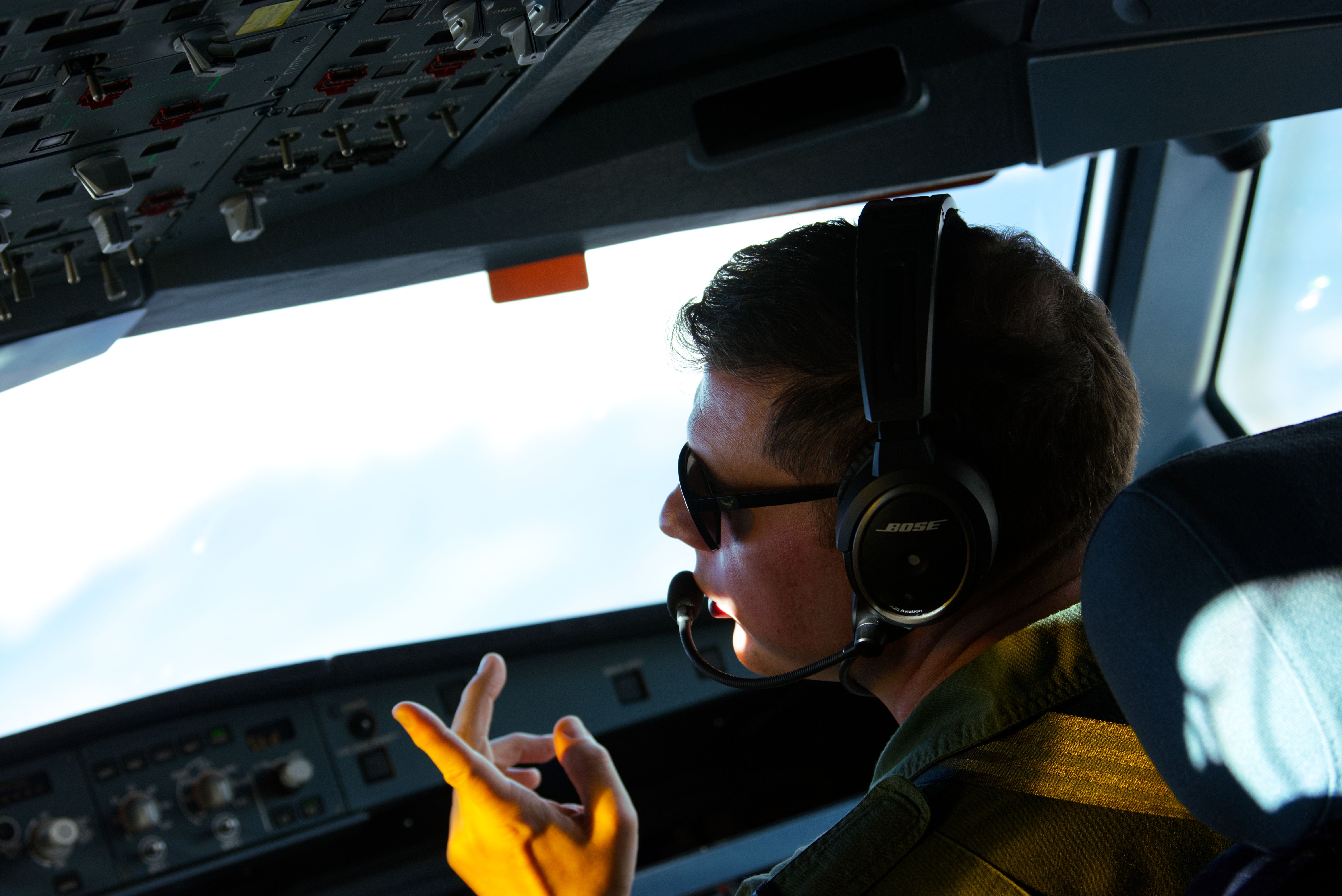 A man wearing sunglasses and Bose headset inside an airplane cockpit, gesturing with his hand. Control panels and lights are visible around him.