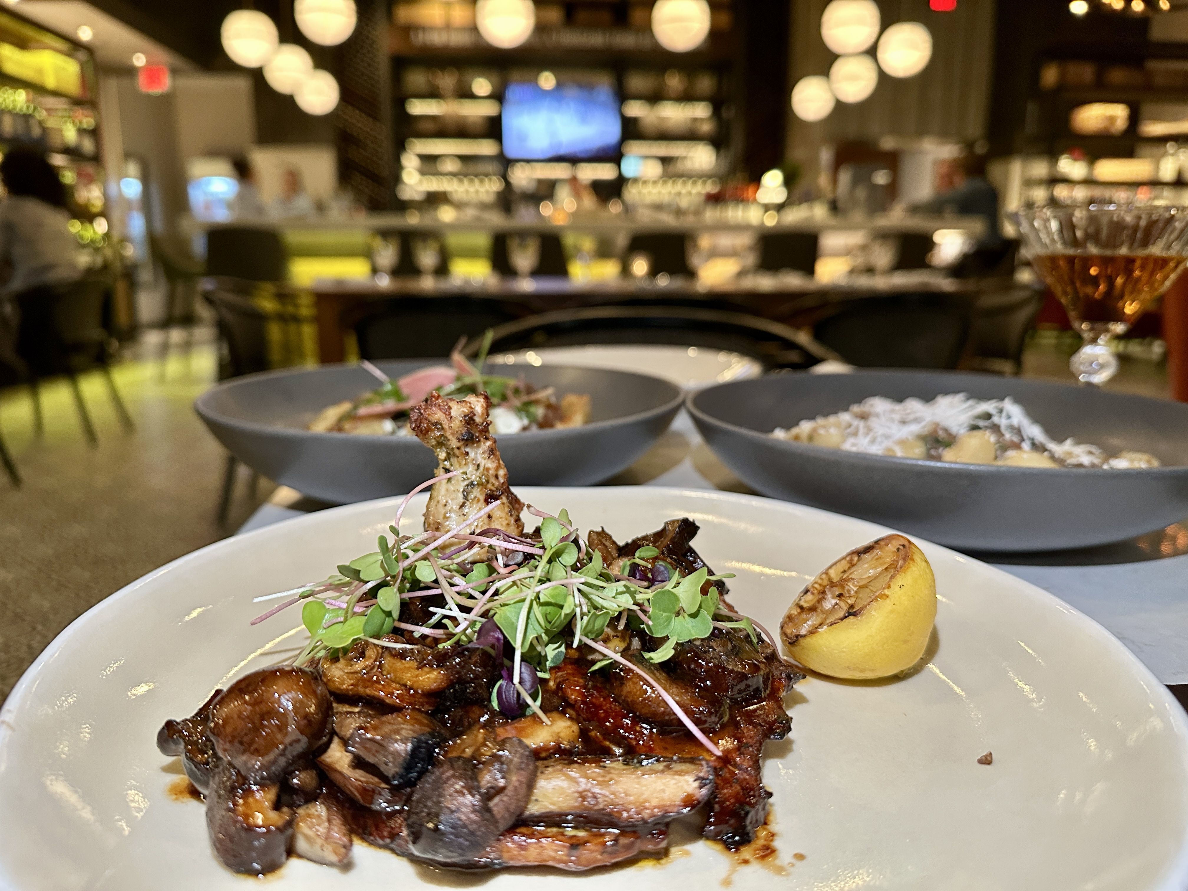 italo uptown charlotte. Close-up of a plated dish with grilled meat topped with microgreens and a lemon wedge, on a white plate; a blurred restaurant bar and warm circular bokeh lights in the background.