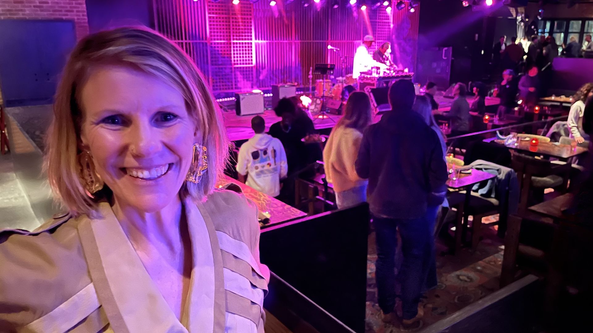 Woman with blond hair smiling standing in front of a stage with a live DJ and neon pink lights.