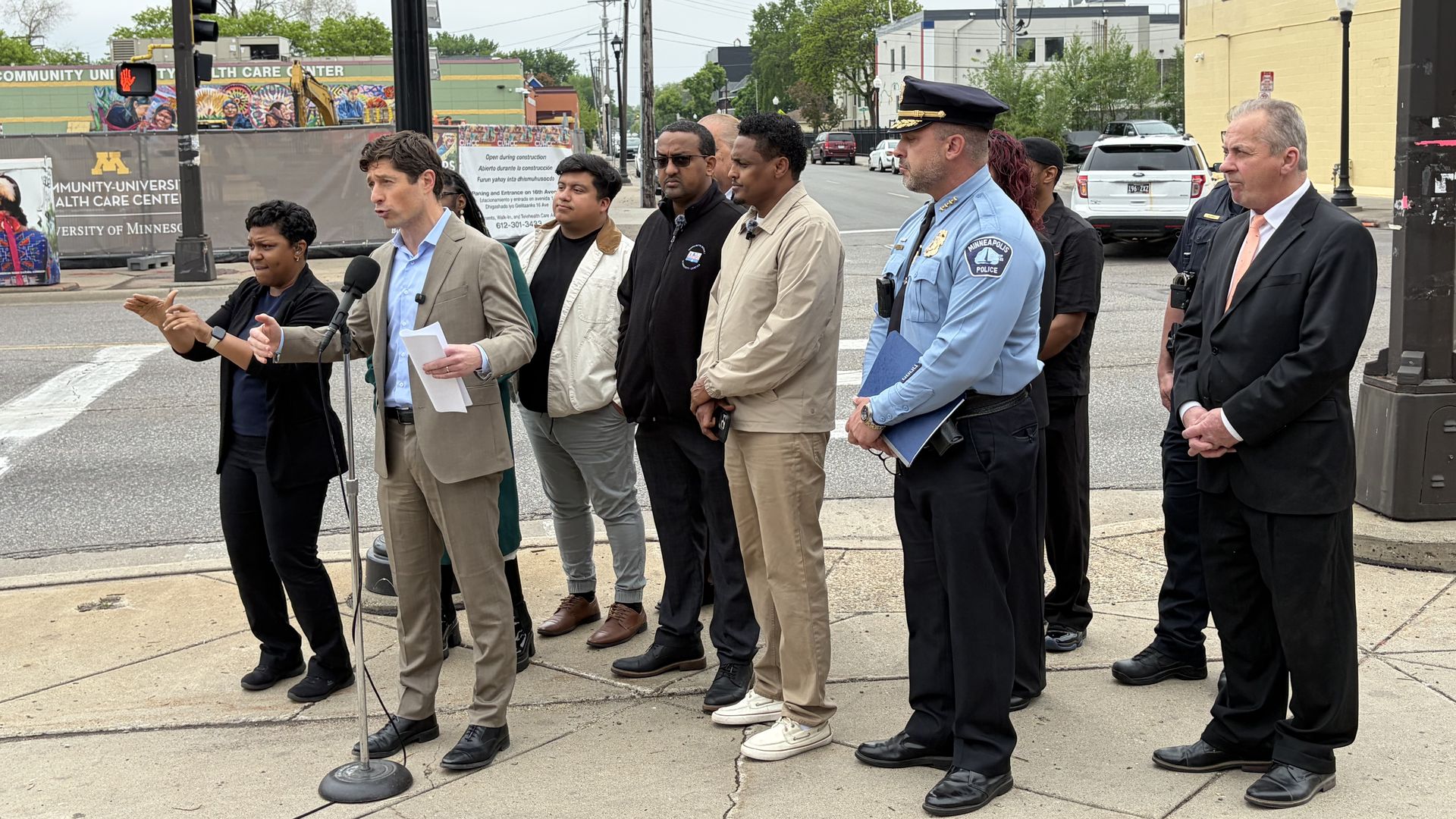 Group of officials and reporters at a street corner press briefing; Mayor Jacob Frey in a suit speaks into a microphone while officers stand nearby, with a colorful mural and city buildings in the background.