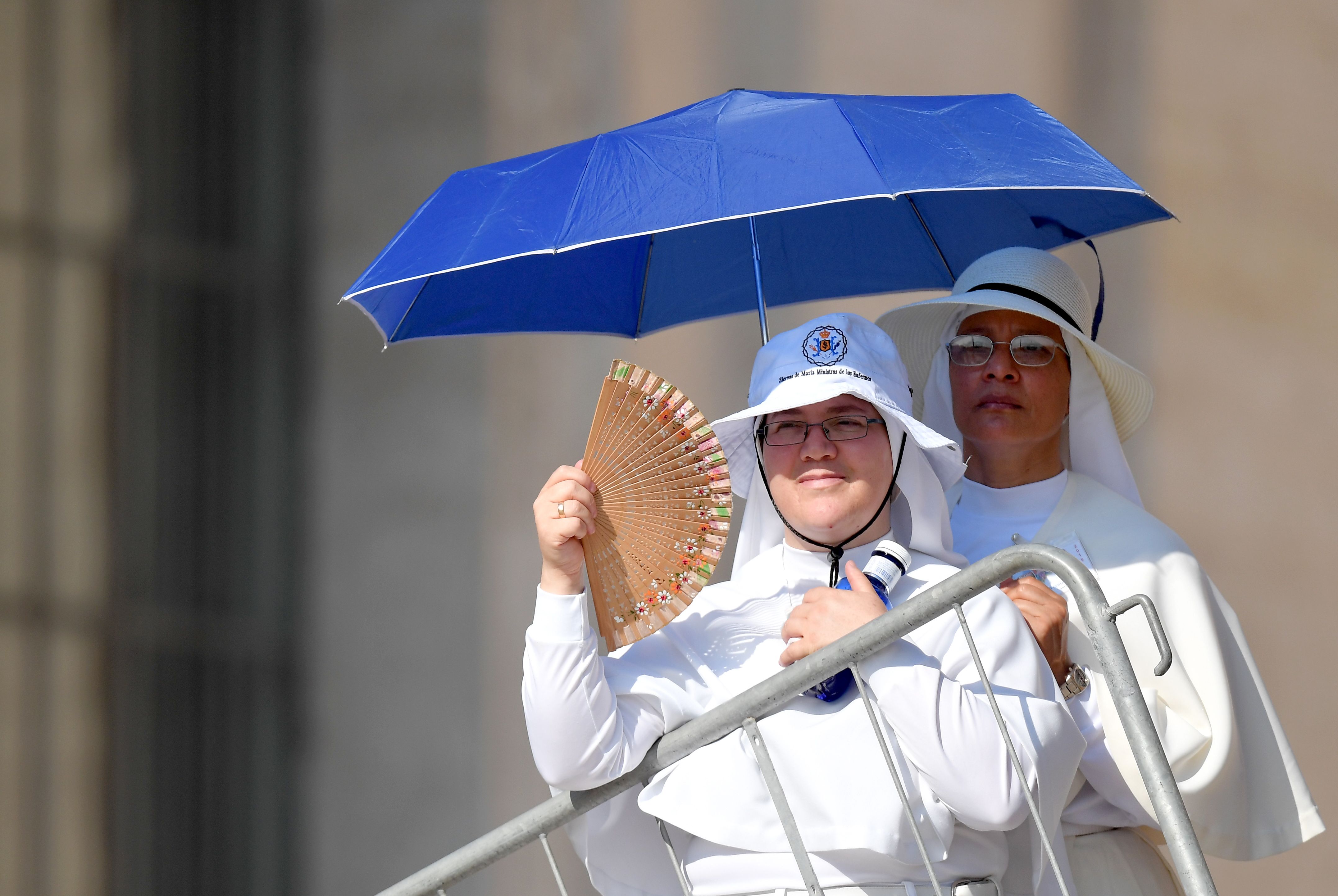 Nuns stand in the shadow of an umbrella and use a fan to protect from the heat on a hot summer day before attending the Pope's weekly general audience at Saint Peter's square at the Vatican.