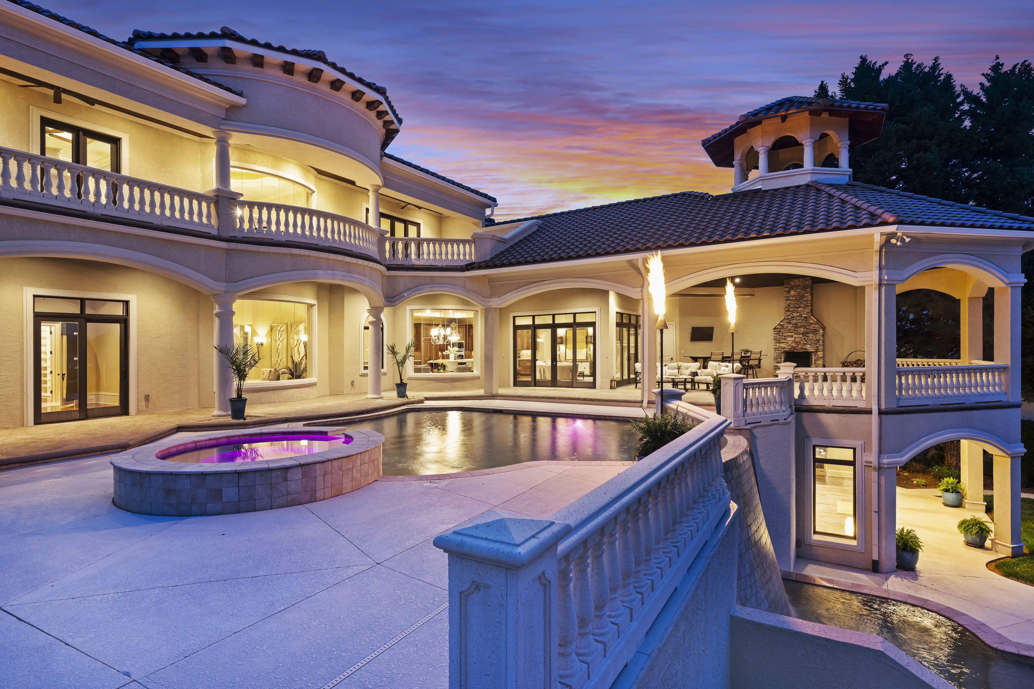 Luxurious Mediterranean-style villa at dusk with curved balconies, columns, and a tiled roof. Outdoor terrace surrounds a pool with purple glow and flaming torches; sunset sky in warm colors.