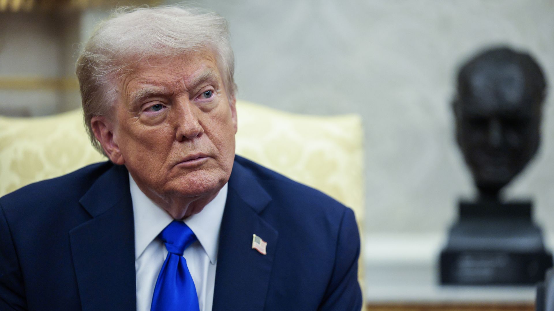 President Donald Trump, wearing a blue suit and sitting in a white and gold chair in front of a white wall and a black bust.