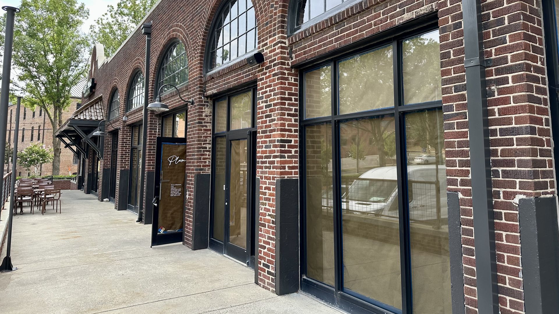 Red brick building with tall arched windows and large shopfront glass along a sidewalk; outdoor seating on the left, green trees, and a bright blue sky in the background.