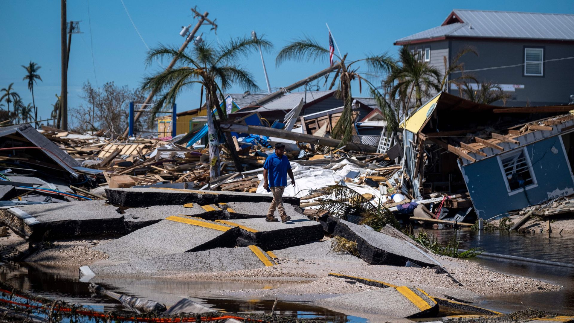A man in a blue shirt and khakis walks on a broken section of road. Behind him are crooked palm trees and debris from toppled houses.