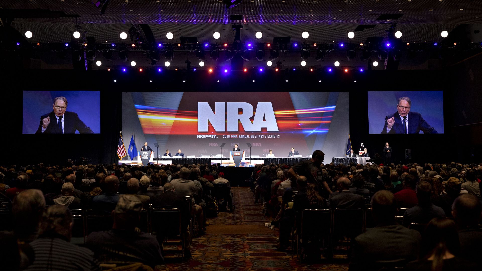 Photo of a person speaking from the stage at a conference with crowds of people and the NRA projected on a screen