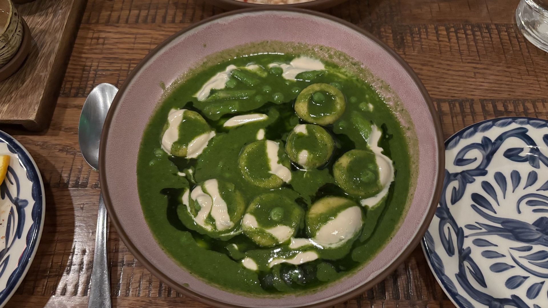 Top-down view of a pink bowl of bright green sauce with round green ravioli, swirls of white cream, on a wooden table beside a spoon and blue-and-white patterned plates.