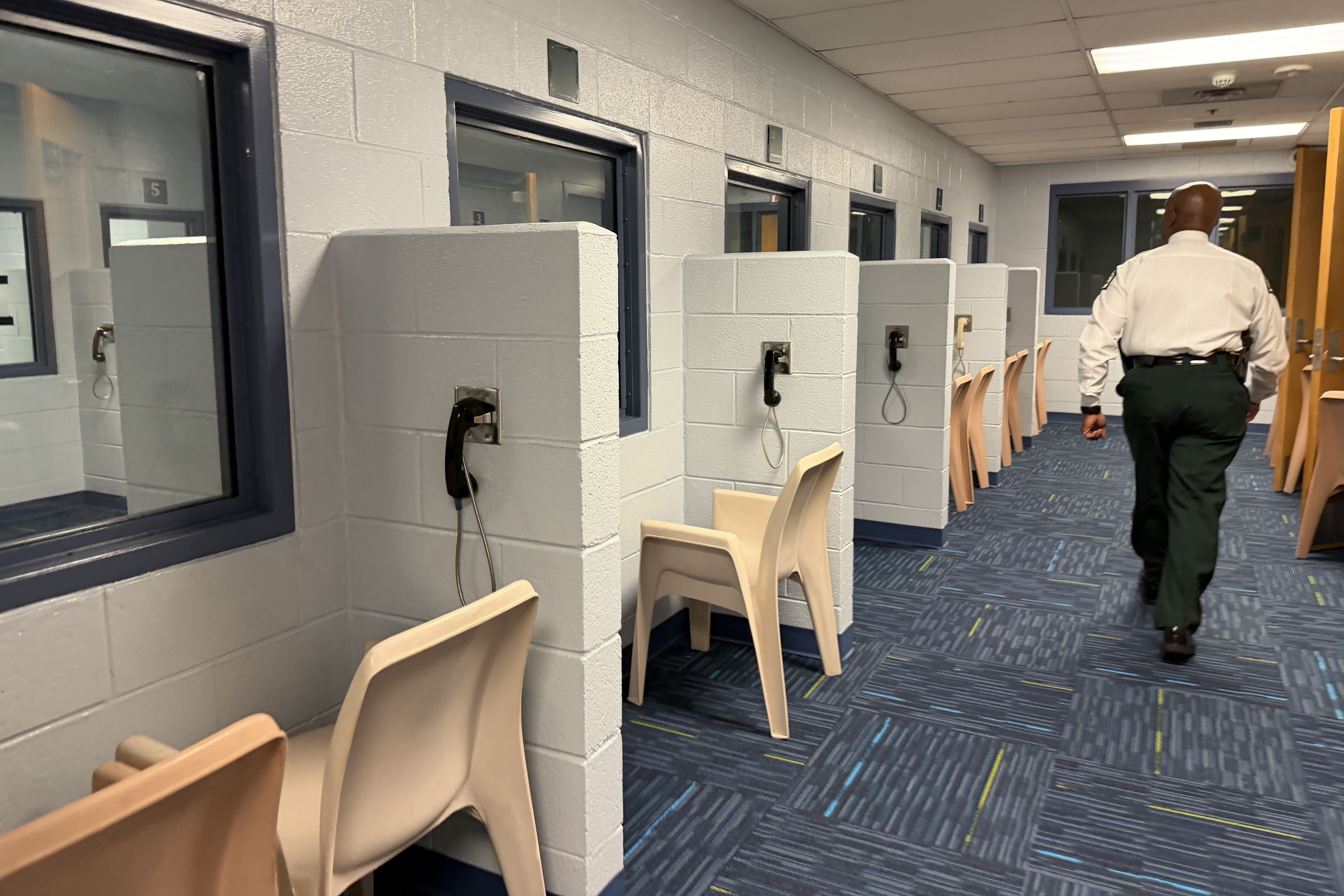 Wide view of a prison hallway with individual cell cubicles along a light gray wall, each with a mounted phone; a security officer in a white shirt walks away toward a doorway at the end.
