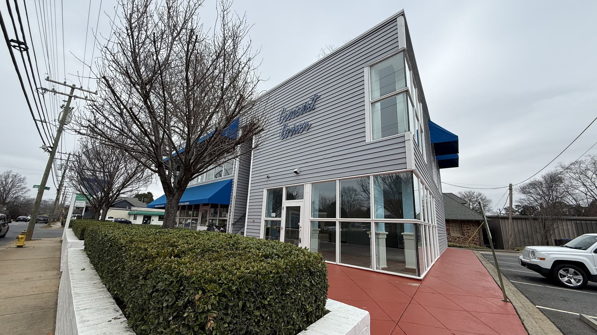 Gray two-story building with blue awnings and signage, red pavement, trimmed green hedges, leafless trees, cars, overcast sky, and a sidewalk with utility poles.