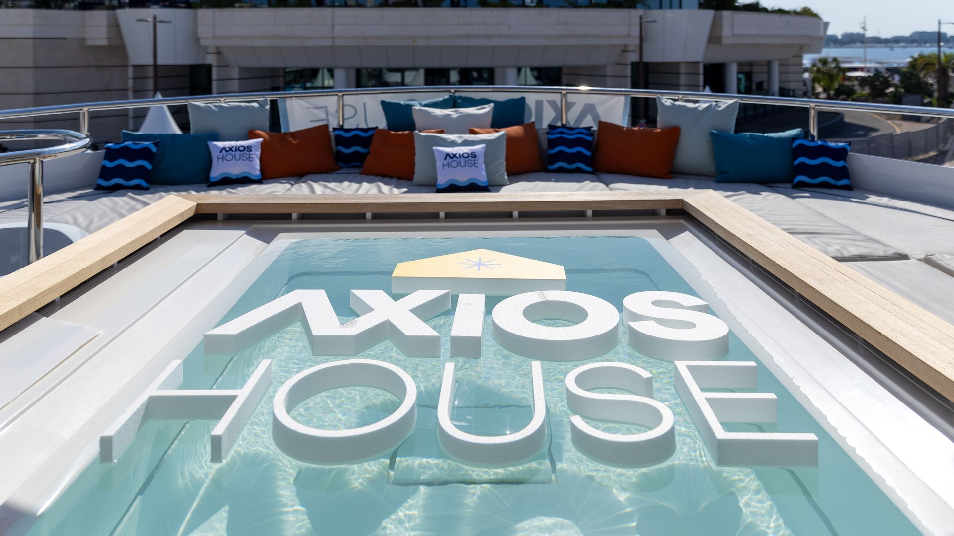 Rooftop lounge with blue, teal, and orange cushions along a white sofa; a glass pool shows large white letters spelling Axios House; a futuristic curved building towers in the background.