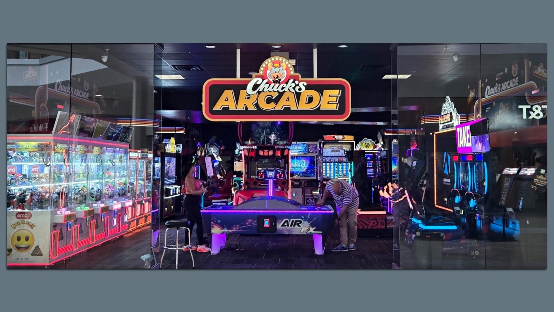 The image shows the entrance of an arcade with a large sign reading "Chuck's Arcade" above it. Inside, various arcade games are visible, including claw machines filled with plush toys on the left. Two people are seen playing an air hockey game in the center. The arcade is decorated with bright, colo