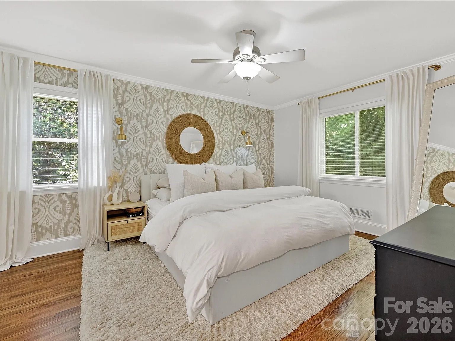Bright bedroom with white bedding, beige patterned accent wall, round wicker mirror, white curtains, wooden floors, a nightstand with decor, ceiling fan, and a large mirror leaning against wall.