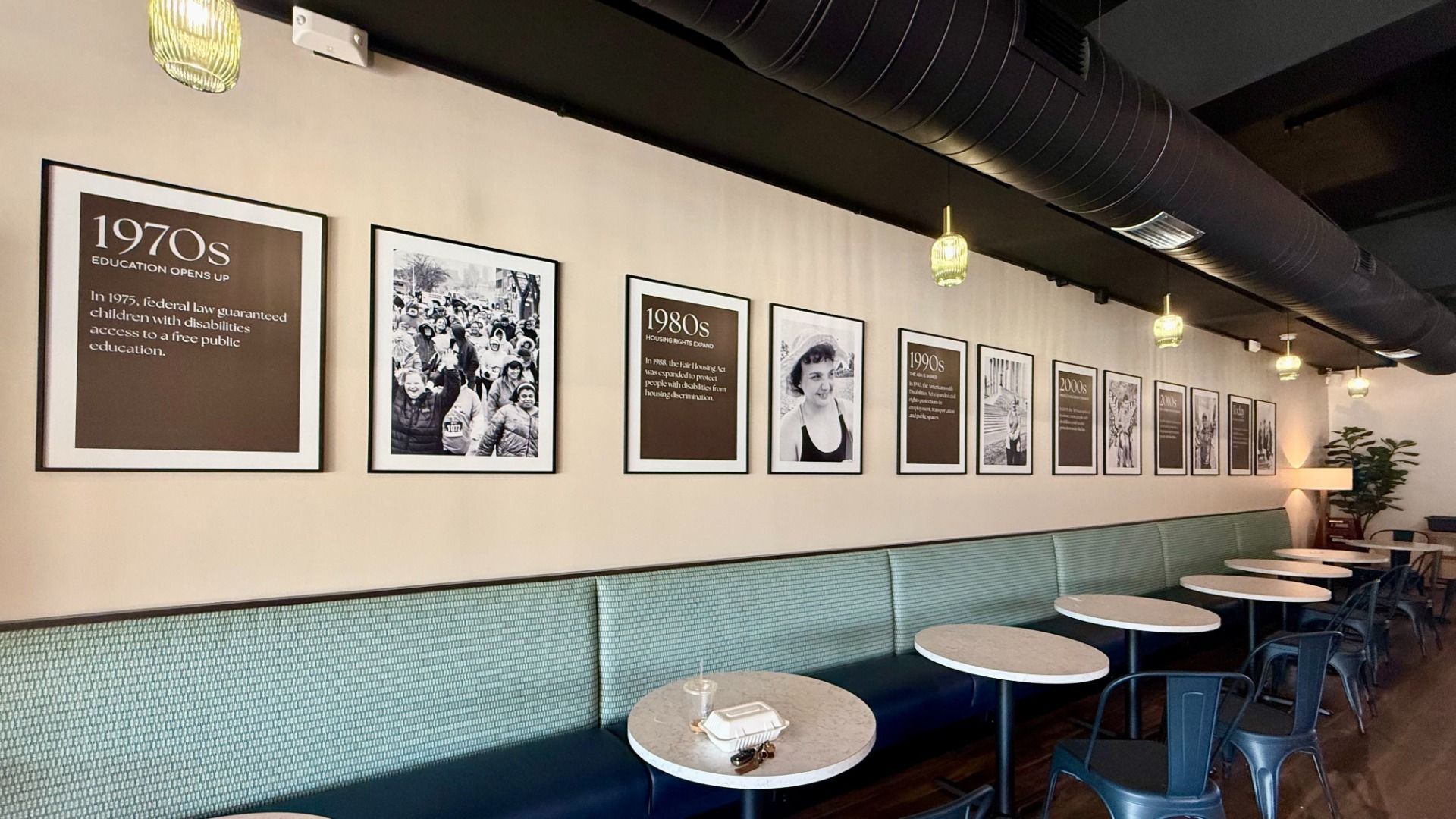 Cafe interior featuring a row of framed panels with 1970s–2000s text and photos, plus a long mint-green bench. Round white tables, blue metal chairs, and yellow-green pendant lights.