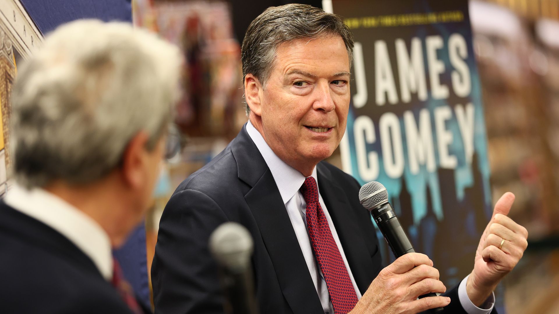 Author James Comey, former FBI Director, in a dark suit and red tie speaks into a handheld microphone at a public event, with a large poster bearing the name James Comey blurred in the background.
