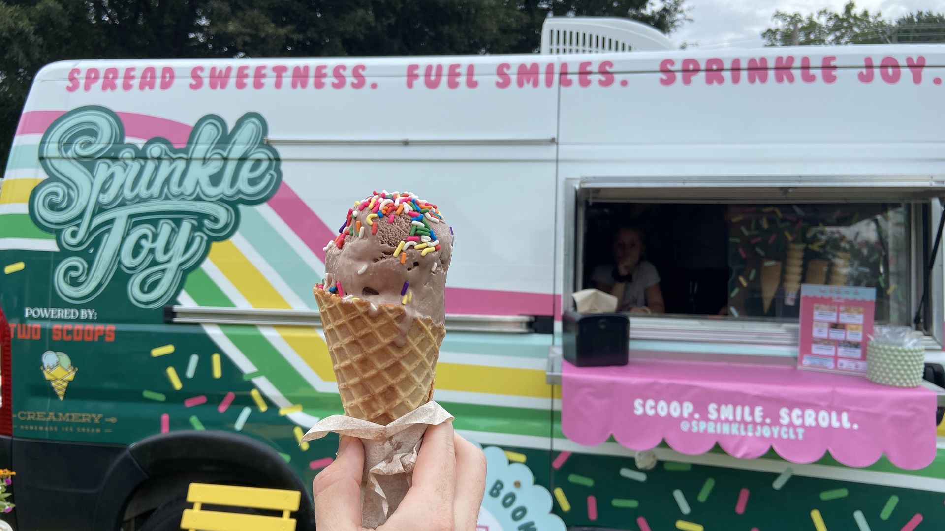 Hand holding a chocolate ice cream cone with colorful sprinkles in front of a pastel-colored Sprinkle Joy ice cream truck with pink, yellow, and green stripes and text.