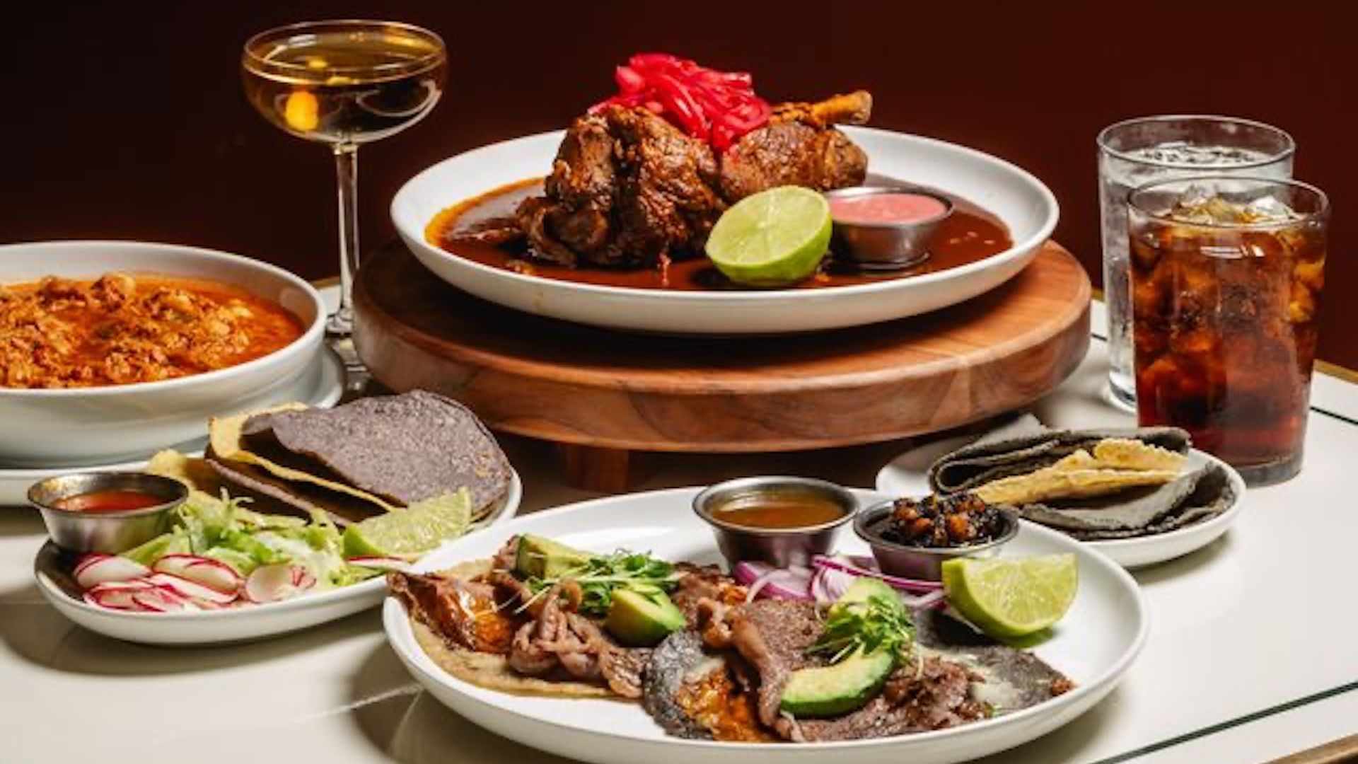 Assorted Mexican dishes on a table: central plate with braised beef, lime wedge and red sauce, surrounded by tortillas, radishes, avocado, a red stew, salsa cups, and a glass of iced drink.
