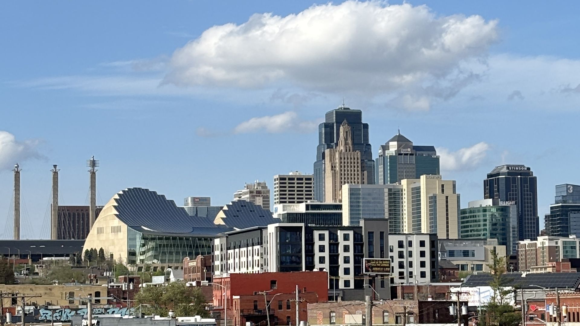 Looking north at the downtown Kansas City skyline with multiple towers, plus the Kauffman Center and the KC Convention Center under a bright blue sky with scattered clouds.