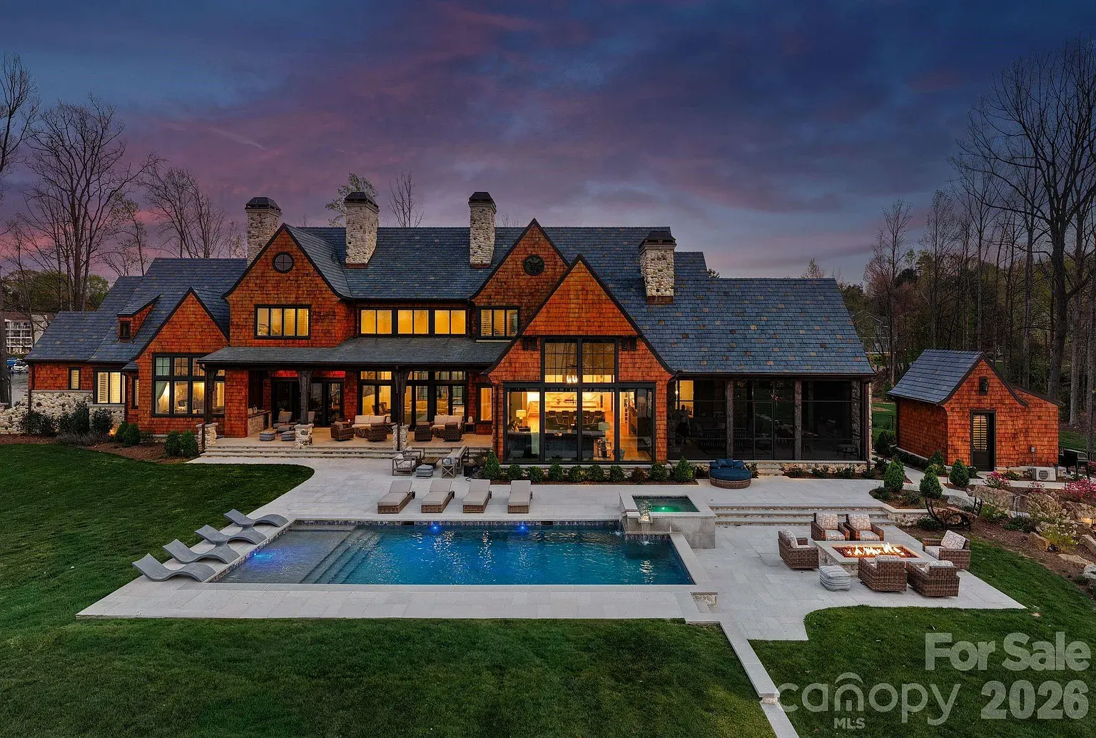 Evening view of a large red-brick luxury home with a slate roof and chimneys. Warm lights glow through the windows; a rectangular pool, lounge chairs, and a fire-pit seating area on a manicured lawn.