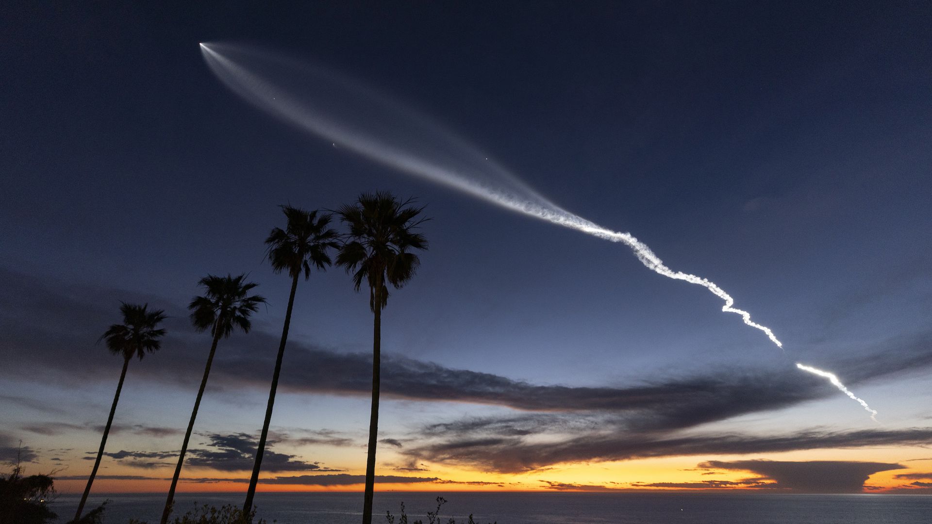 A rocket soars across the night sky over the ocean at sunset.